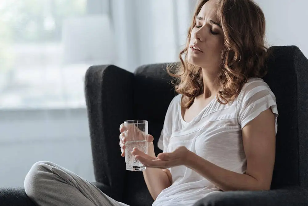 a woman sitting in a chair holding a glass of water about to take a pill