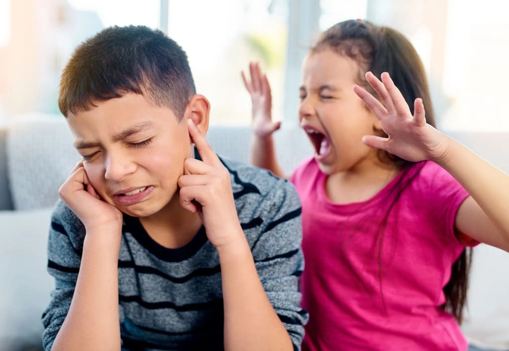 a little girl screaming next to a boy that covers his ears