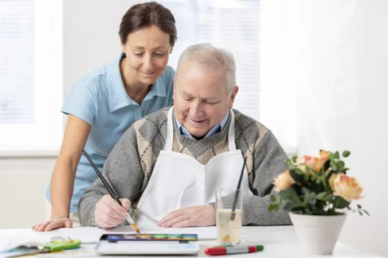 a female support worker watching an old man painting on a table at home